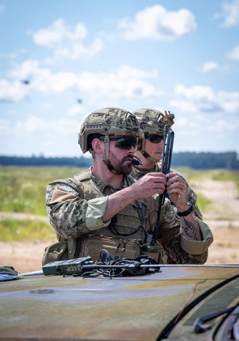 Two Canadian Armed Forces members in camouflage uniforms and helmets prepare communications equipment beside a military vehicle during a training exercise at Sēlija Range, Latvia. An open training area stretches into the distance beneath a bright blue sky dotted with white clouds.