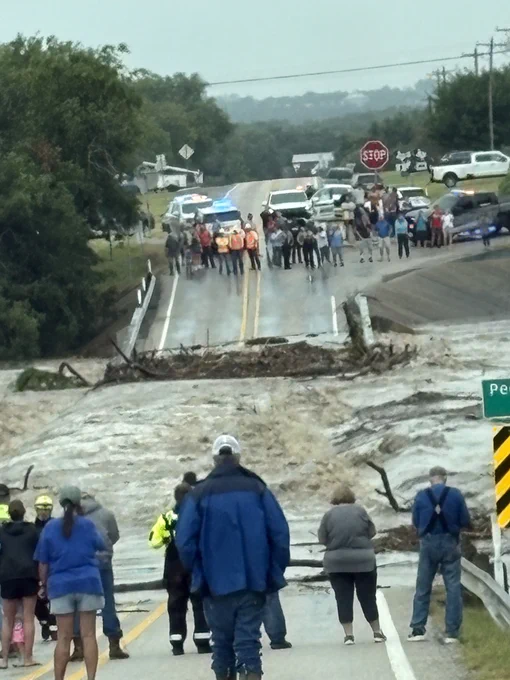 A picture of RM 1623 across the Pedernales River after the heavy rain began. There's heavy water flowing across the roadway with people standing on both sides of the roadway.
