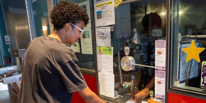A person stands at a colorful ticket counter, placing a hand on the metal surface. The counter has various posters and information. A star-shaped decoration is visible on the glass.