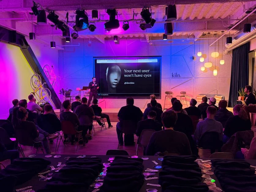 A speaker presents to an audience at a meetup, discussing app design for AI users, with a projector displaying relevant text.