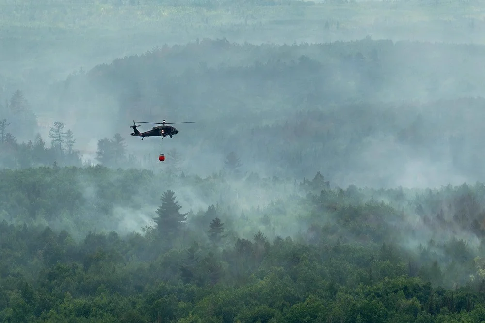 A helicopter flies above woodlands engulfed in smoke and haze. 