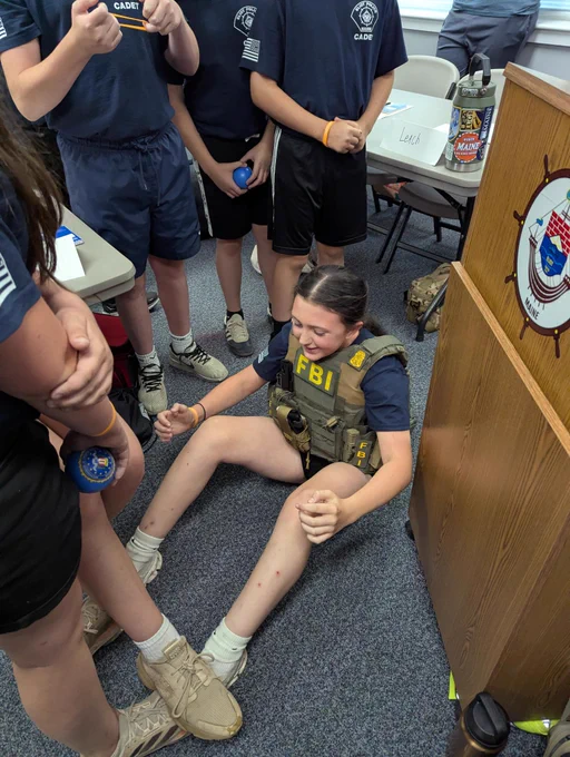 Image is a group photo taken during an FBI Boston visit to the Eliot Police Department Youth Academy in Eliot, Maine. Photo shows can academy member wearing an FBI tactical vest while doing sit-ups during an exercise. 