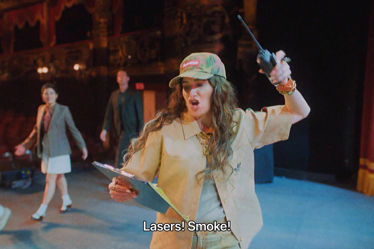 From The Studio. Kathryn Hahn as Maya Mason is yelling on stage holding a clipboard in one hand and a walkie talkie in the other. Text on the bottom of the screen reads "Laser! Smoke!"