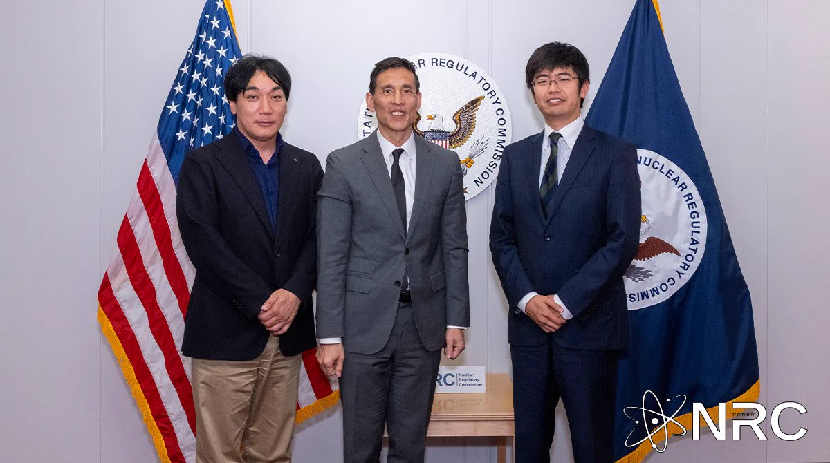 Image is a photo of three people standing in front of the NRC flag and seal and the American flag, smiling at the camera.