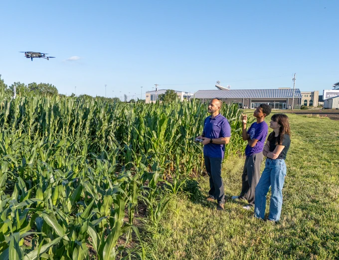 Three individuals standing in a field of tall corn, observing a flying drone. One person is holding a remote control. Several buildings are visible in the background, including one with a sign reading "Walmart.