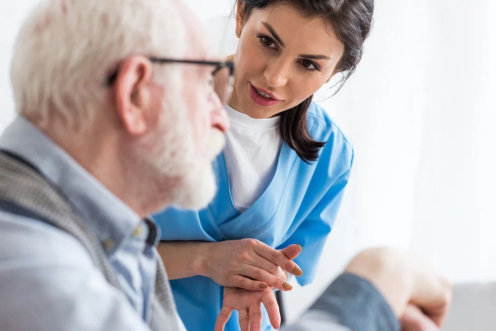 A woman in a medical uniform speaks with a seated older man.