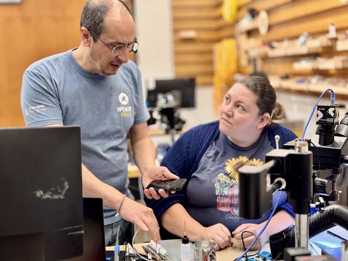 An instructor demonstrates smartphone repair techniques to a student at a microsoldering workstation. The instructor holds a phone. The bench is equipped with a stereo microscope, soldering tools, flux, and electronics repair equipment.