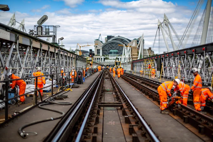 Engineers working on the Hungerford Bridge. On left staff in organge are repainting the steelwork. On the right operatives work on track. In the distance work in the platforms can be seen.