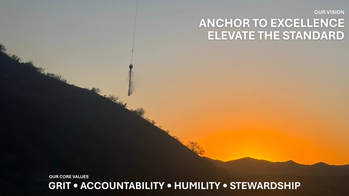 A helicopter drops a bucket of water on a wildfire as the sun is setting. The text reads: our vision, anchor to excellence, elevate the standard. And the core values of grit, accountability, humility and stewardship.