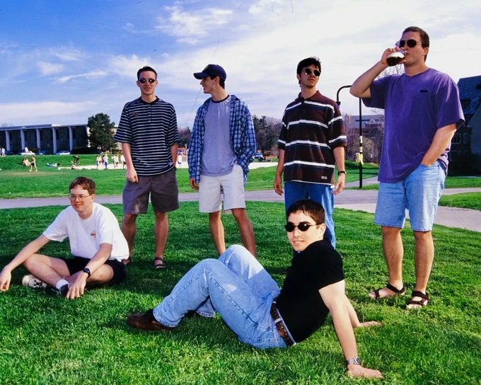 A group of Cornell students casually enjoy time on a grassy field. Some are sitting, while others stand. They are dressed in casual attire, including sunglasses and T-shirts. In the background, people are scattered across the green, and a large building is visible.