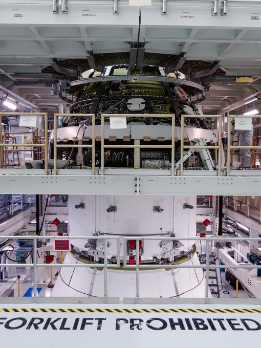 The Orion crew and service modules for NASA’s Artemis III mission are shown mated in a workstation in the Operations and Checkout Building at the agency's Kennedy Space Center. The view shows some of the crew module’s exposed wiring and structural elements on the upper section and the white exterior of the service module below, all surrounded by work platforms, railings, signs, and facility hardware. Credit: NASA