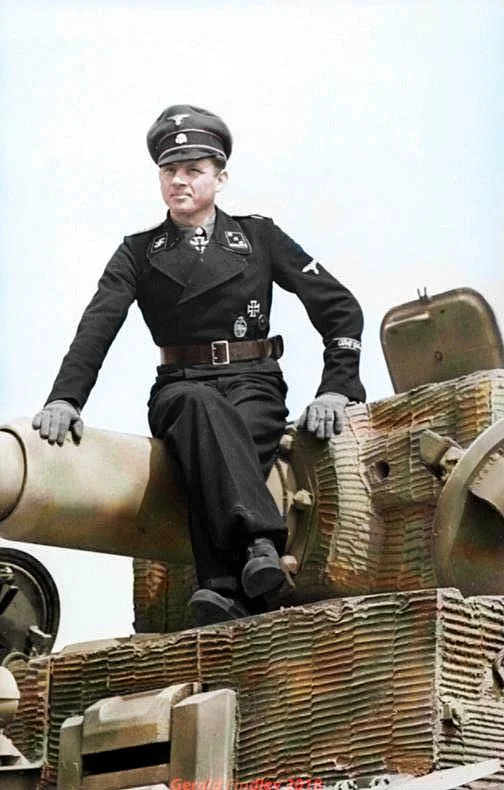 VoicesofWW2's tweet photo. On the morning of June 13, 1944, a single German tank rolled into the flank of a British armored column outside a Norman town called Villers-Bocage and, in about fifteen minutes, tore the head off an entire elite division. The man commanding it was Michael Wittmann, and it is probably the most famous single tank action in history.

Wittmann was already a celebrated tank ace by 1944, a veteran of the Eastern Front where he had run up a huge score of Soviet tanks knocked out, and he wore the Knight's Cross with Oak Leaves. In Normandy he commanded a company of Tiger tanks, the heavy, thickly armored German tanks with the long, deadly 88 millimeter gun that could kill an enemy tank from far beyond the range at which they could hurt him.

That morning the British 7th Armoured Division, the famous Desert Rats, were driving to seize Villers-Bocage and push behind the German line. A long column of their tanks and armored vehicles halted along the road, crews relaxed, some brewing tea, unaware that Tigers were nearby. Wittmann, in a single Tiger, saw the strung-out column and did not wait for support. He attacked it alone.

He rolled along the flank of the stationary British column and destroyed it methodically, one vehicle after another. He knocked out the tanks at the front and the rear to trap the rest on the road, then worked down the line, brewing up Cromwell tanks, Sherman Fireflies, scout tanks, and half-tracks that could not maneuver or bring their guns to bear in time. In roughly a quarter of an hour his one tank destroyed something on the order of a dozen or more tanks plus around fifteen personnel carriers and a couple of anti-tank guns, before his Tiger was finally disabled and he escaped on foot.

His attack shattered the British thrust and helped throw the whole operation into disorder, costing the Desert Rats heavily in men and machines. It made Wittmann a national hero in Germany. He was killed in his Tiger less than two months later, in August 1944, during the fighting as the Normandy front collapsed. But the fifteen minutes at Villers-Bocage put him permanently into the history of armored warfare.