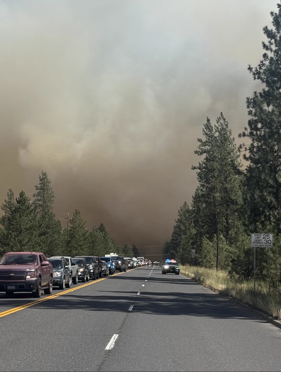 Smoke billows in the distance. On the left, a line of cars waits to safely evacuate. On the right, a cop car is parked with their emergency lights on and the rest of the roadway remains clear for emergency responders using the lanes to travel westbound towards the fires.