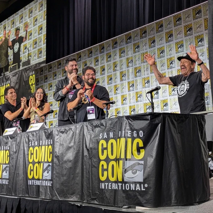 A panel at San Diego Comic-Con International. Danny Trejo stands at the microphone with both arms raised in celebration, wearing a Trejo's Tacos t-shirt and cap. Four panelists seated at the black Comic-Con branded table applaud and smile beside him, with a large screen and Comic-Con step-and-repeat backdrop behind them.