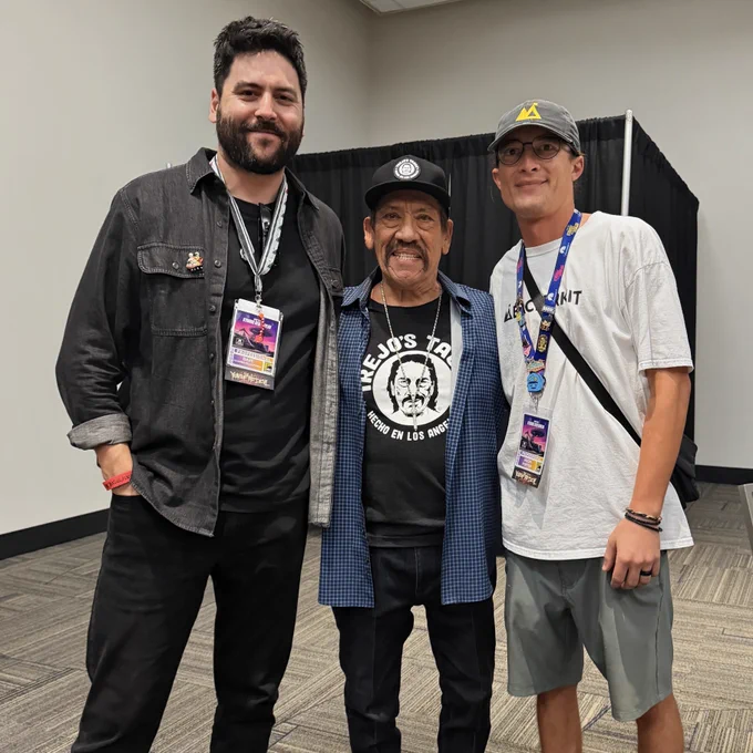 three men standing together and smiling in front of a black curtain at San Diego Comic-Con. Danny Trejo is in the center wearing a Trejo's Tacos t-shirt, cap, and blue plaid shirt, flanked by two men wearing professional convention badges, one in a dark denim shirt and one in a white BackerKit t-shirt and cap.