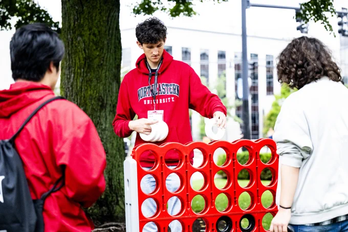 A young man in a red 'NORTHEASTERN UNIVERSITY' hoodie places a white disc into a large red Connect Four game. Two people stand nearby: one in a red jacket with a black backpack, and another with curly hair in a light top, observing the outdoor game.