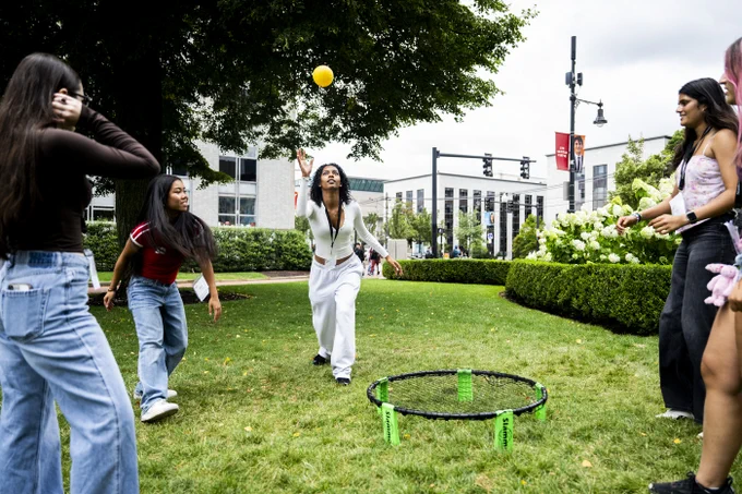 Diverse young women play spikeball on a vibrant green campus lawn. One in white reaches for a yellow ball above the Slammo net; others watch.