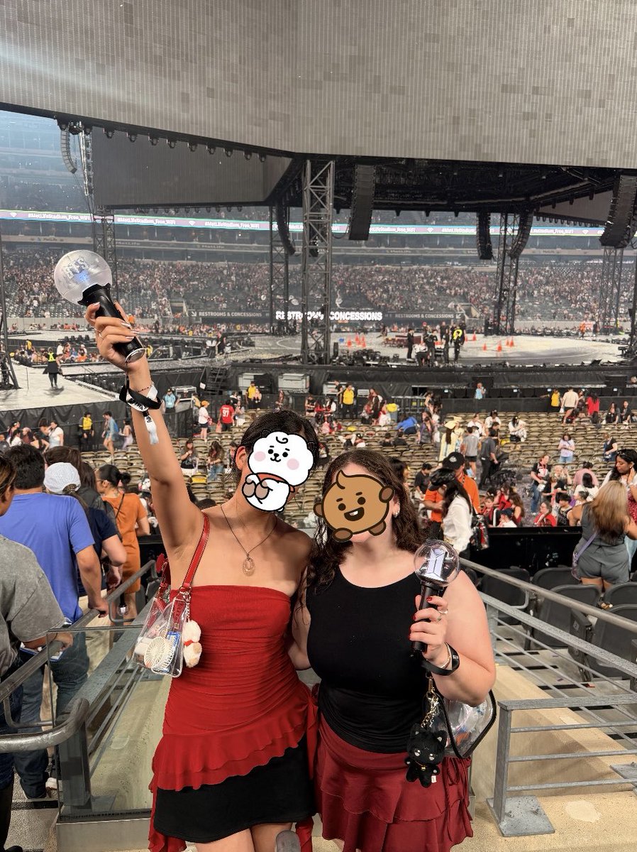 Two girls posing with their BTS lightsticks in front of a concert stage.