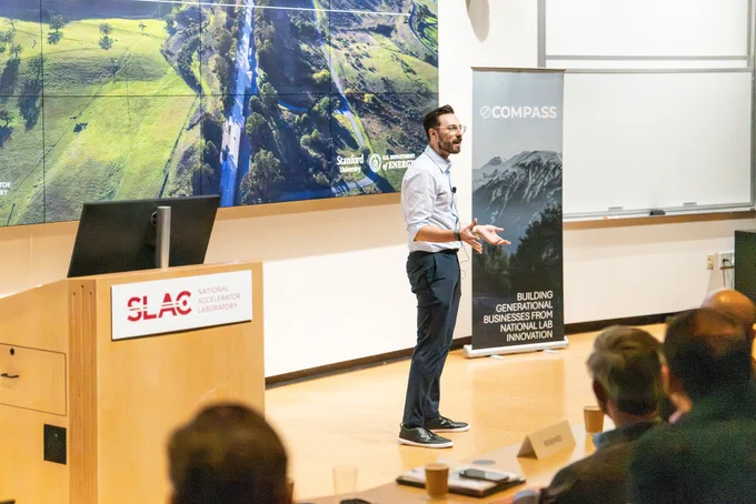 A person stands giving a presentation in a lecture room. Behind them is a large screen showing a scenic landscape, with logos of Stanford University, SLAC National Accelerator Laboratory, and COMPASS. A banner reads, "Building Generational Businesses from National Lab Innovation." Audience members are seated in the foreground.