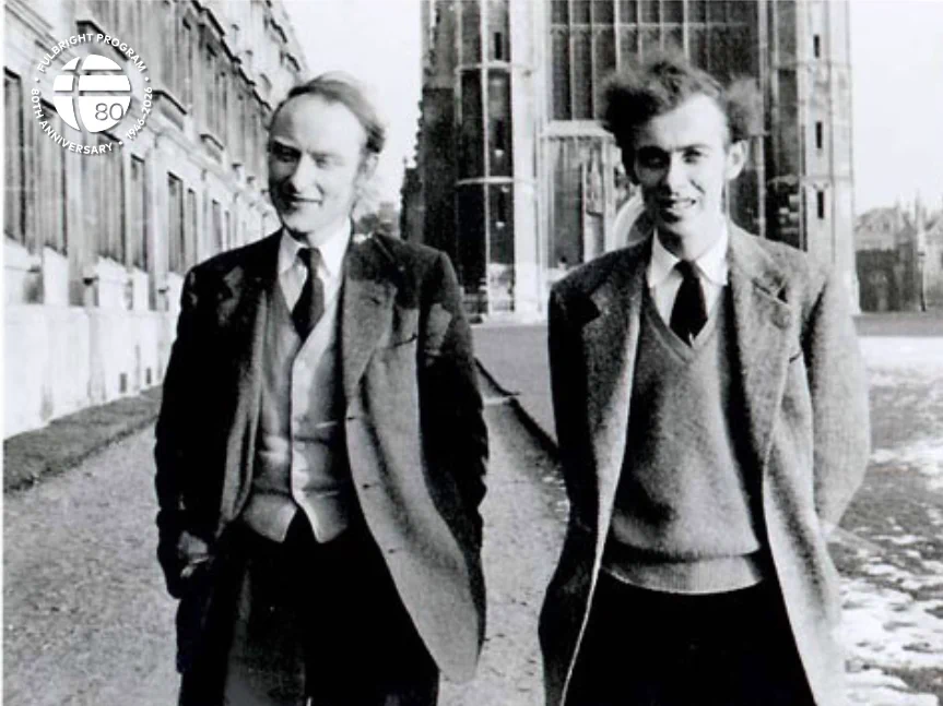 Two individuals walking on a path in front of a large building. Both are wearing suits and are outdoors. A university emblem is visible in the top-left corner of the image.