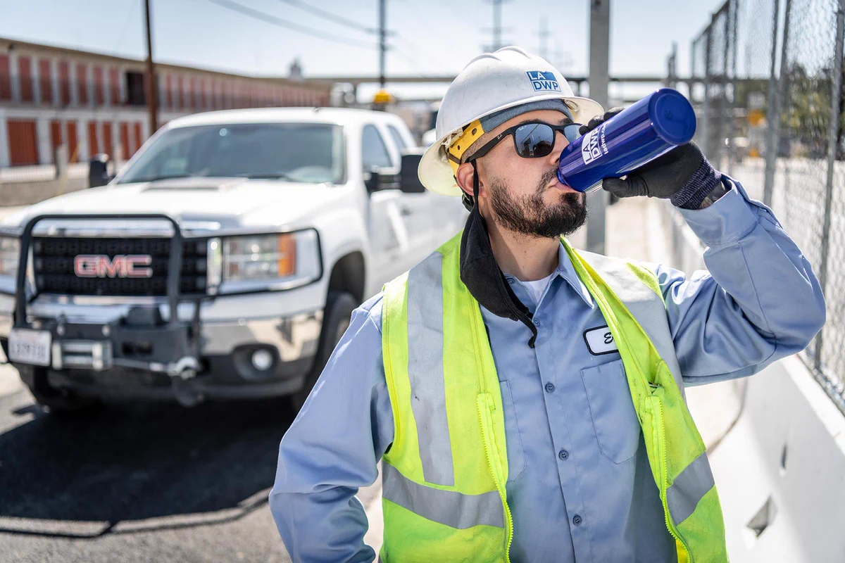 A man in LADWP safety gear and sunglasses standing in front of a white truck drinking from a water bottle.
