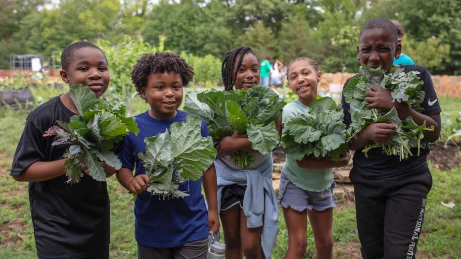 A group of children stand in a garden, each holding leafy green vegetables. The background features various plants and a few people working in the garden. The setting appears lively and natural.