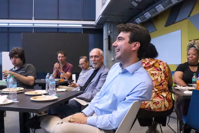 A sitting man in a blue shirt laughing. He is surrounded by others at a lunch in the Coda Building.
