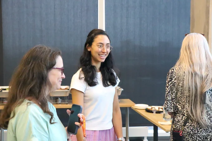 A smiling woman in glasses speaks to someone out of frame of the camera. There is a woman on either side of her.