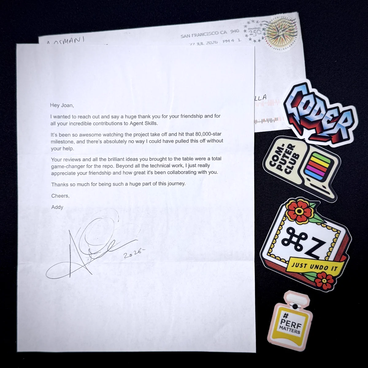 A printed thank-you letter from Addy Osmani on a dark background, hand-signed and dated 2026, next to its opened envelope postmarked San Francisco. Four stickers lie beside it: "Coder", "Computer Club", a Cmd+Z one reading "Just Undo It", and a perfume bottle reading "#perfmatters".