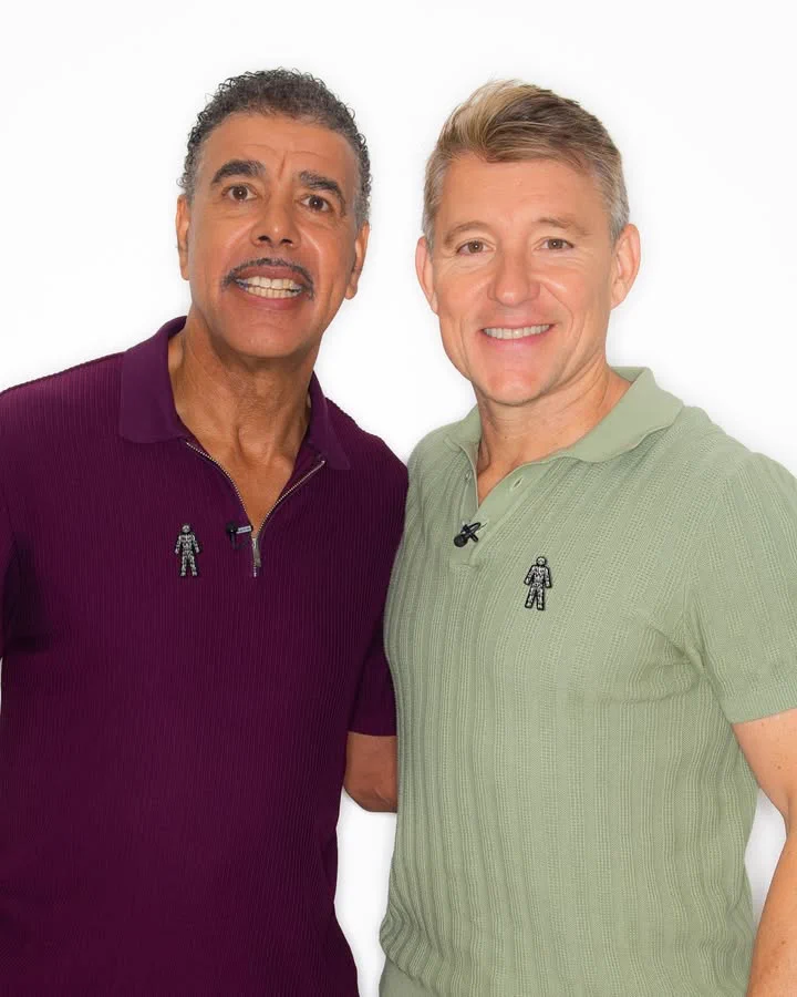 Chris Kamara and Ben Shephard standing together, smiling at the camera. They are both wearing polo shirts with a Prostate Cancer UK Man of Men pin badge attached.