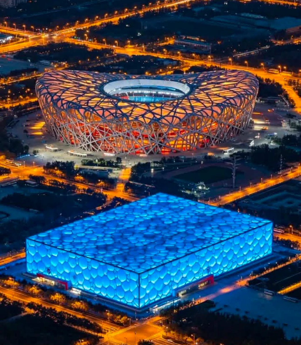 xb5808's tweet photo. As night falls, the Bird's Nest glows with warm structural lights while the Water Cube shimmers in cool blue. Iconic Beijing Olympic landmarks shine brightly amid city street lights.
 #BeijingBirdsNest #WaterCube #BeijingOlympicPark https://t.co/VNRnxlpGid