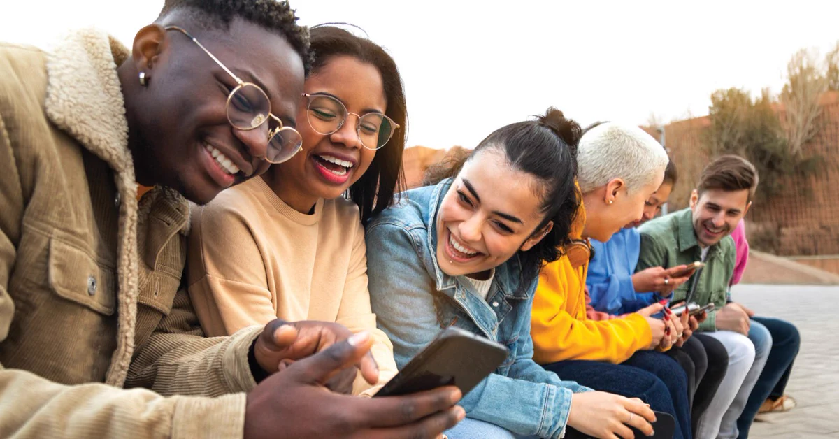 A group of people sitting outdoors on a bench, looking at a smartphone with smiles. They are casually dressed and seem engaged with each other. It's a bright day.