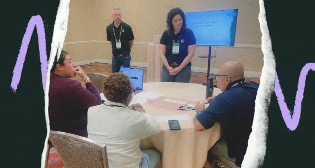 A group of three individuals at a round table engaged in discussion, with a fourth person standing in the background beside a presentation screen in a conference room.
