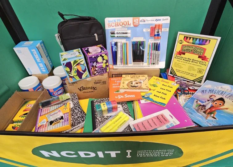 A box filled with various school supplies, including notebooks, pens, markers, folders, glue sticks, and tissue boxes. There are books like "My Magical Choices" and "One Fish Two Fish Red Fish Blue Fish" by Dr. Seuss. The front of the box features the NCDIT logo.