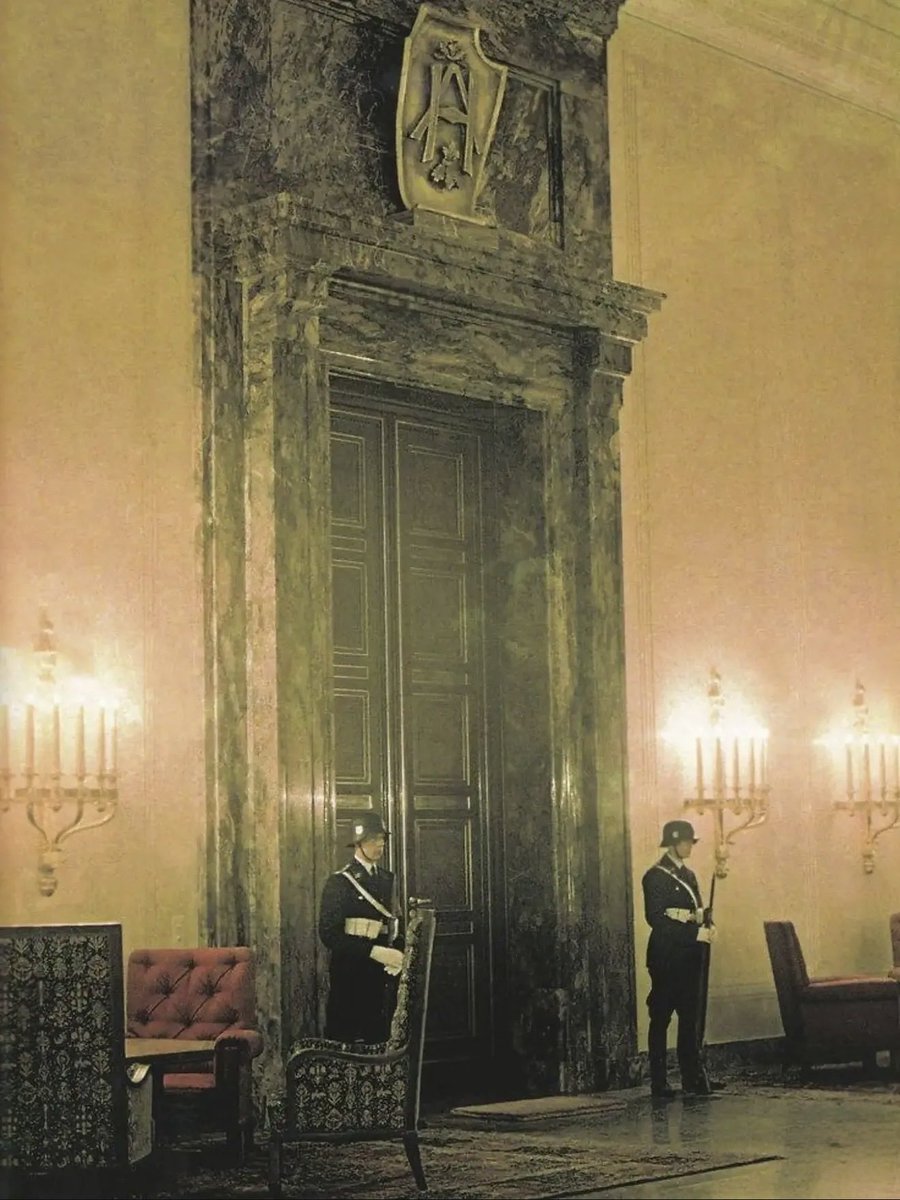 NeSS4201889's tweet photo. The entrance to Adolf Hitler's personal office within the New Reich Chancellery in Berlin. The room was designed by Albert Speer and featured imposing, oversized architecture intended to intimidate visitors. The doorway is framed by dark marble and topped with a shield bearing a stylized eagle emblem. Guards from the Leibstandarte SS are positioned on either side of the entrance.