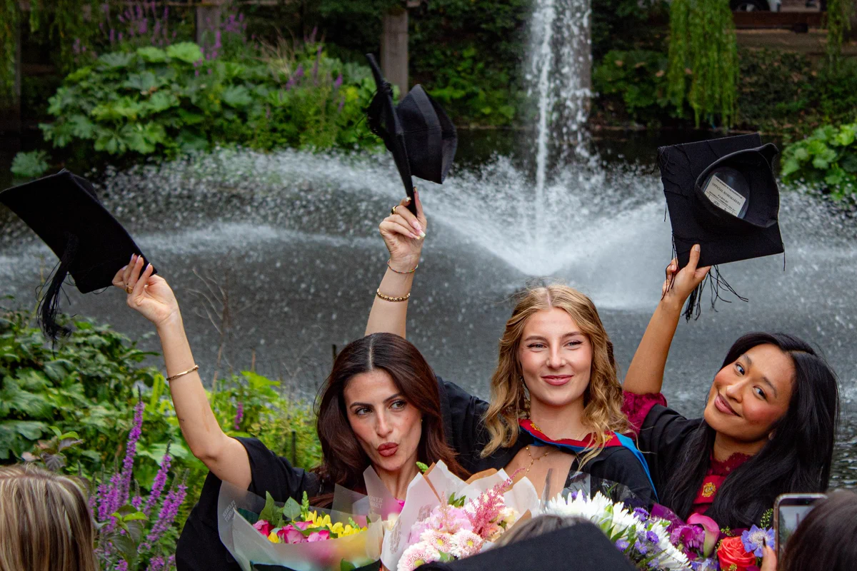 Three graduates hold their caps in the air. All three are holding bouquets in their other hand.