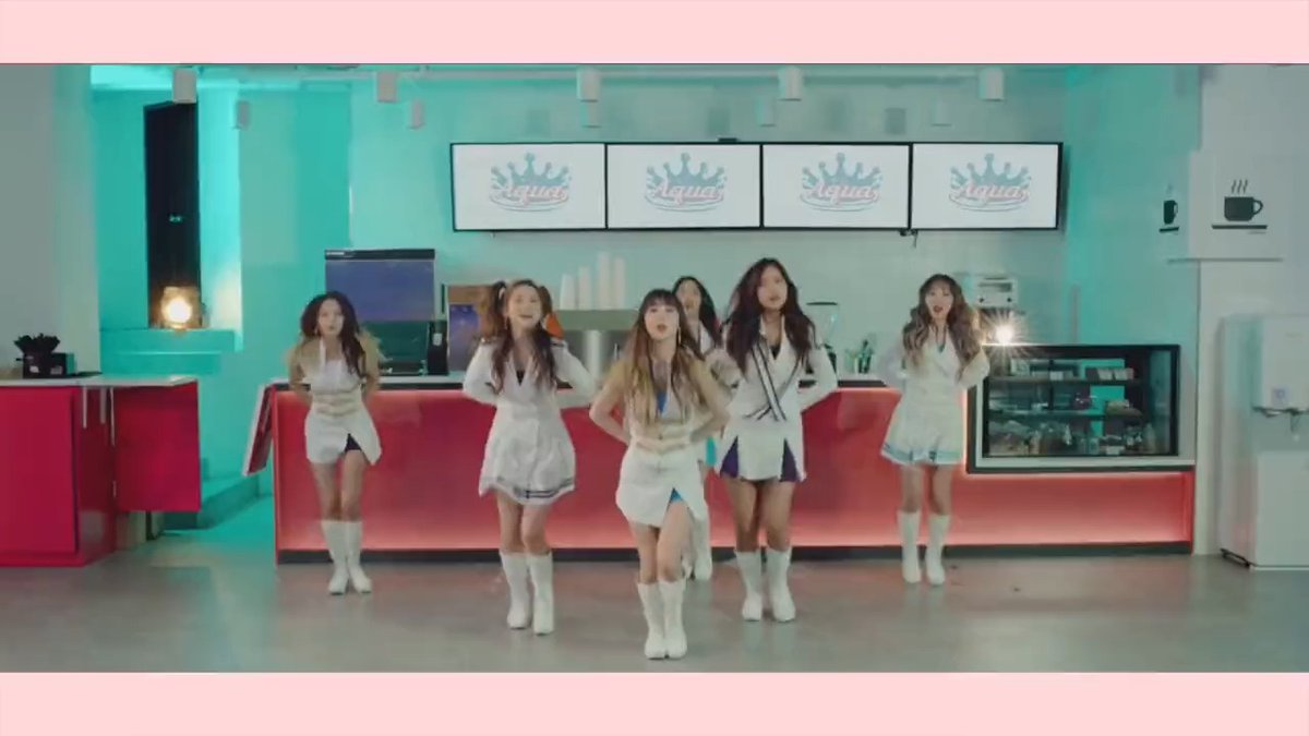 Six women in coordinated white and cream outfits with white boots sing and dance in a brightly colored set featuring teal walls and pink accents, with "Aqua" logos on screens behind them.