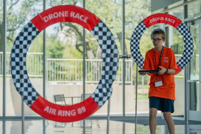 A student in an orange shirt operates a drone through a circular obstacle labeled "Drone Race Air Ring 89" indoors.