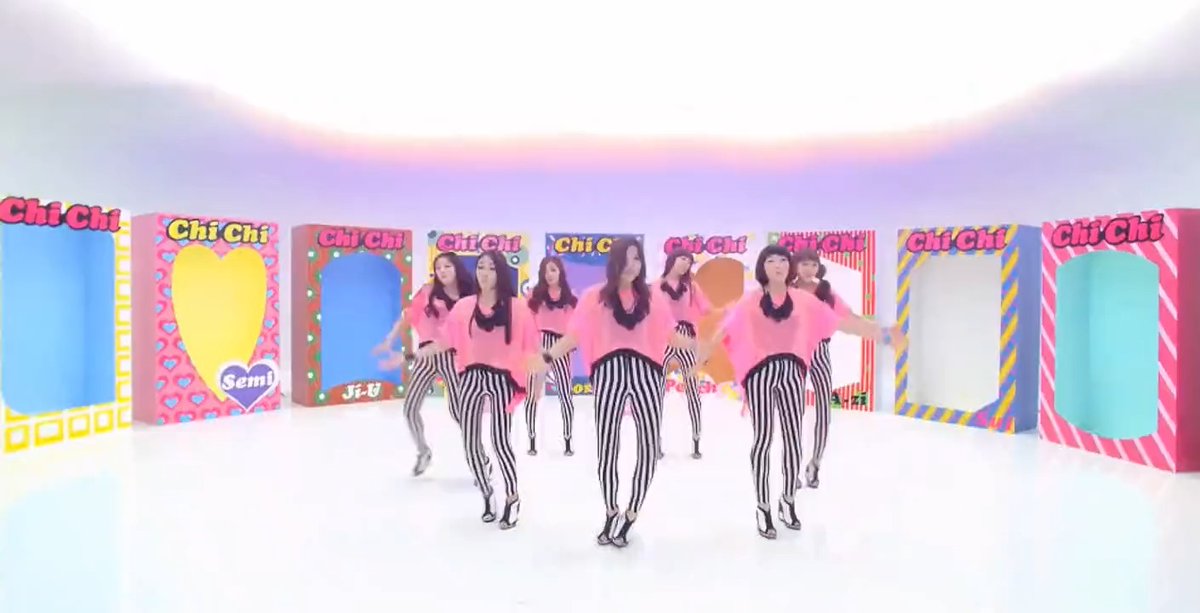 Several identical women wearing pink tops and black-and-white striped pants dance in a colorful studio setting surrounded by display boxes labeled 'Chi Chi'.