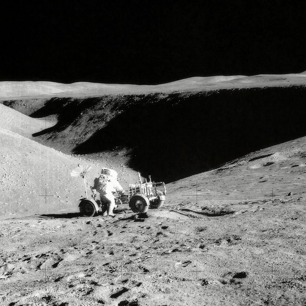 universetheta's tweet photo. That black band running across the middle is not a shadow on a hillside. It is a hole in the ground, and the far side of it is about a kilometer and a half away.

This is the Moon, on 31 July 1971. The man at the vehicle is David R. Scott, commander of Apollo 15. The photograph was taken by James B. Irwin from the flank of a nearby crater, looking north along a valley called Hadley Rille.

The valley is roughly 1.5 kilometers wide and about 400 meters deep, and it winds for more than 100 kilometers along the foot of a mountain range. The sun is high and the sky behind it is completely black, because there is no air up there to scatter the light.

Look at the small crosses scattered across the picture. Those are not damage. There is a glass plate inside the camera with a grid etched into it, so that every frame comes back with a built-in ruler and any distortion in the film can be measured and corrected afterward.

Now look at where they parked, because the parking spot is the experiment.

Almost everywhere on the Moon the solid rock is buried. Billions of years of impacts have ground the top of it into loose powder and rubble, meters deep. If you want the rock underneath you have to dig for it, and they had hand tools and a drill and a few hours.

This valley is a cut that was already made. Lava flowed along here and carved down through the plain, and the walls it left are a vertical slice through the ground. The upper sixty meters or so of that wall expose layers of basalt, individual lava flows stacked one on another, most of them at least ten meters thick.

So they did not dig down. They drove to the edge of the one place where the ground was already open, and photographed sideways into it with a telephoto lens.

One honest note. That lava carved this valley is agreed. Whether it ran as an open channel in the open air, or ran inside a roofed tube that later fell in, is still argued over.

The vehicle is the reason any of this happened. It was carried to the Moon folded up so that the crew could get several kilometers away from the lander instead of a few hundred meters. Without it, this valley was simply out of reach.

Image: NASA. Hasselblad camera on the lunar surface, Apollo 15, edge of Hadley Rille, 31 July 1971. Photograph by James B. Irwin. Further cropped and sharpened.