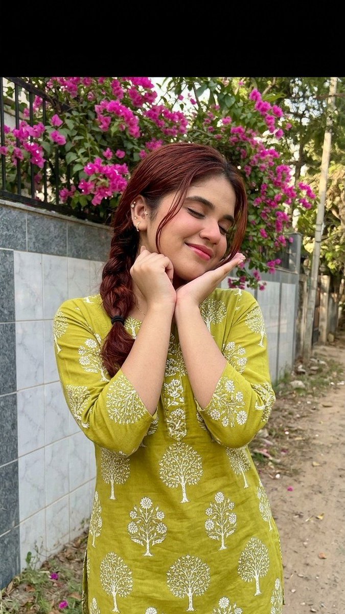 oye_samia's tweet photo. Feeling as bright and cheerful as these blooms!🌼

Prompt: Hyper-realistic full-body portrait of a young South Asian girl outdoors. Wearing lime/olive-green cotton kurti with white tree/floral block print, three-quarter sleeves, dark blue jeans. Long black hair in loose side braid with side-swept strands. Small stud earrings, natural minimal makeup, soft pink lips. Playful pose with hands under chin, head tilted, eyes closed, smiling. Background: boundary wall with grey-white tiles, vibrant pink bougainvillea, dirt path, natural daylight. Don’t change her face.
