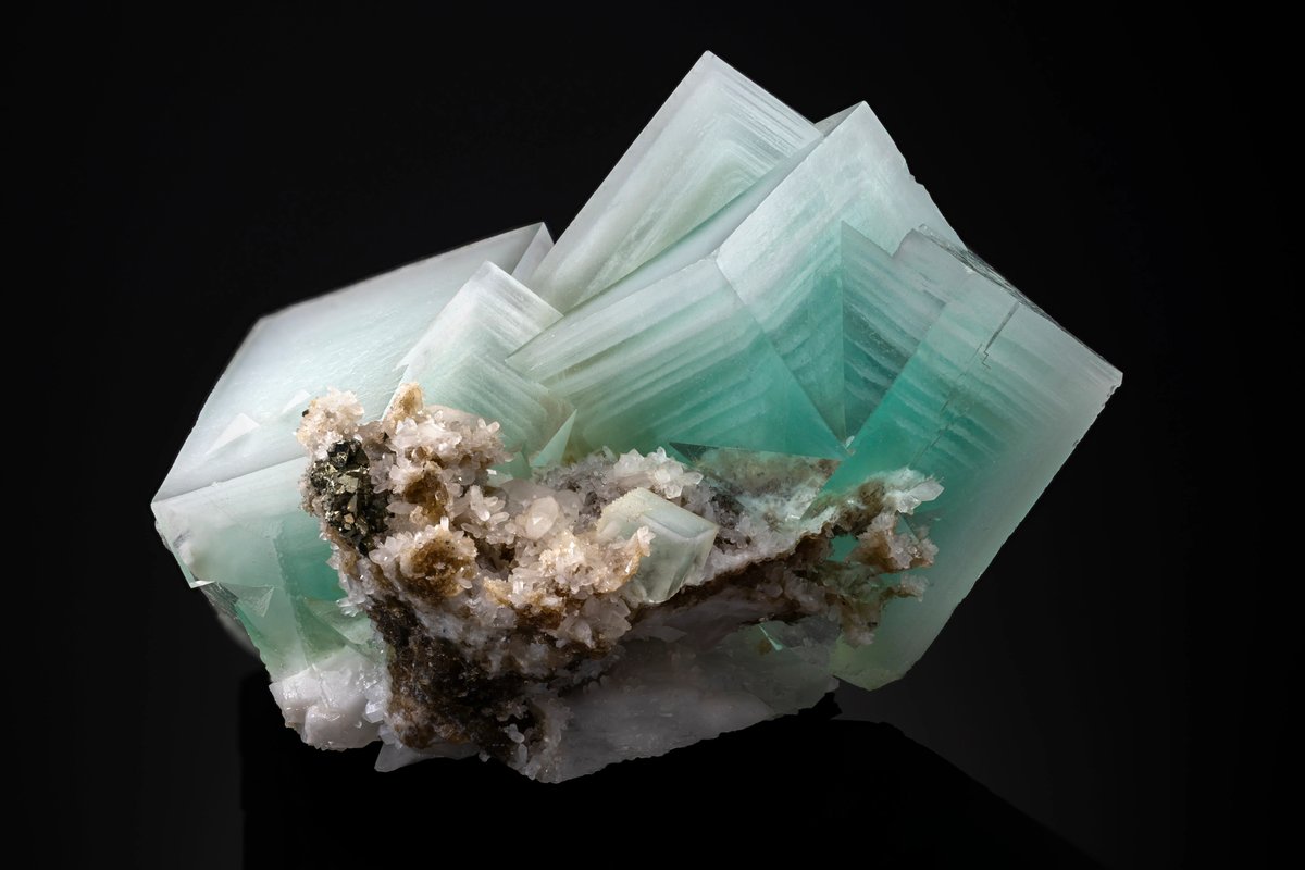 RockHoundRick's tweet photo. Been turning this Yaogangxian piece over in my head all afternoon because the growth habit on it is just plain weird for fluorite. Normal fluorite gives you clean cubes or interpenetrant octahedra, flat faces, done. This thing grew like a pagoda roof, stair stepped layers stacking back from each face instead of one smooth plane. You can actually count the ledges on the pyramidal terminations like tree rings turned sideways.

What's happening on the atomic level is that the crystal wasn't growing at a steady rate. Each face was building forward in pulses, and every time growth outran the supply of calcium and fluorine ions in the surrounding fluid, the leading edge stalled just slightly before the next pulse of saturated solution shoved growth forward again. Do that dozens of times and instead of one flat face you get a terraced stack, basically a fossilized record of the pocket chemistry lurching between undersaturated and oversaturated.

What gets me is the locality. Yaogangxian throws some of the sharpest, most photogenic cubic fluorite on the planet, that's the mine's whole reputation, crisp edges and saturated color zoning. So a pocket that also produced this stepped habit means the fluid there was capable of two totally different growth regimes depending on how stable conditions stayed during crystallization. Steady flow gives you the textbook cubes everyone collects Yaogangxian for. Turbulent, pulsing flow with temperature or pressure hiccups apparently gives you this.

I keep picturing what the pocket actually looked like while this was forming, fluid pushing in, dropping its load, backing off, pushing in again, over some span of time nobody can pin down exactly. Rare habit, and it makes me wonder how many other stepped specimens got broken down at the mine before someone realized what they were holding.

Link in 🧵