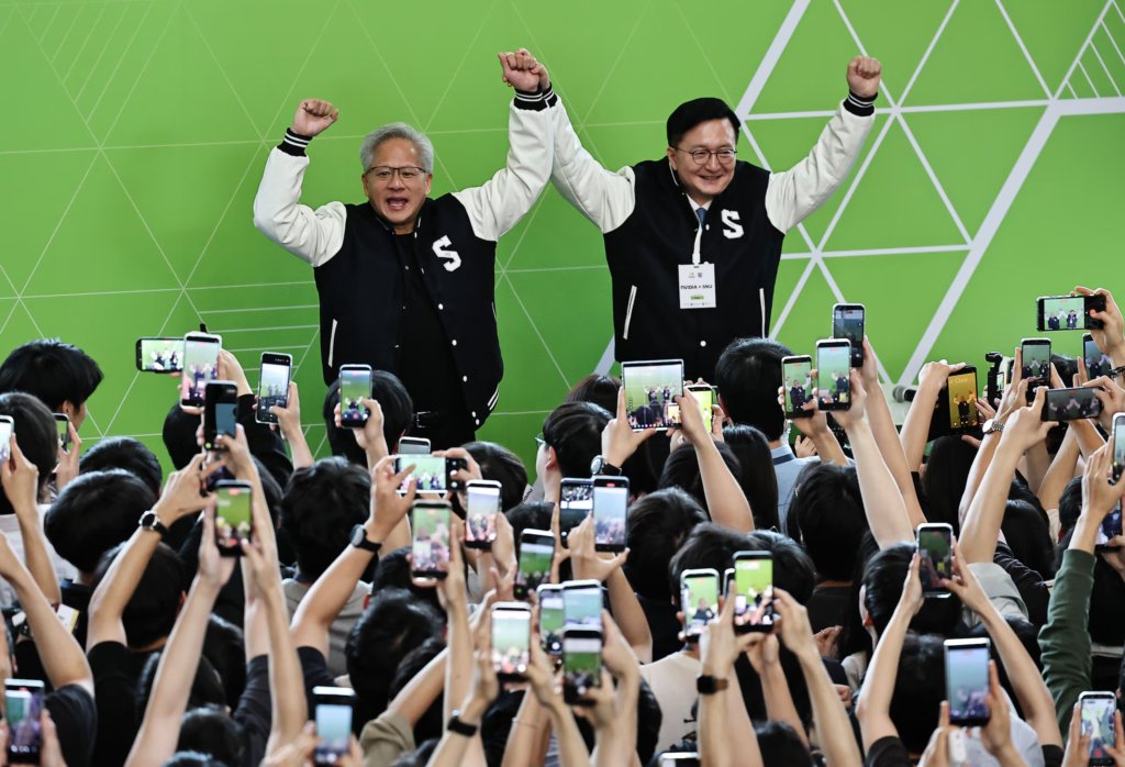 Two people on stage raising their arms as a crowd takes photos with phones.