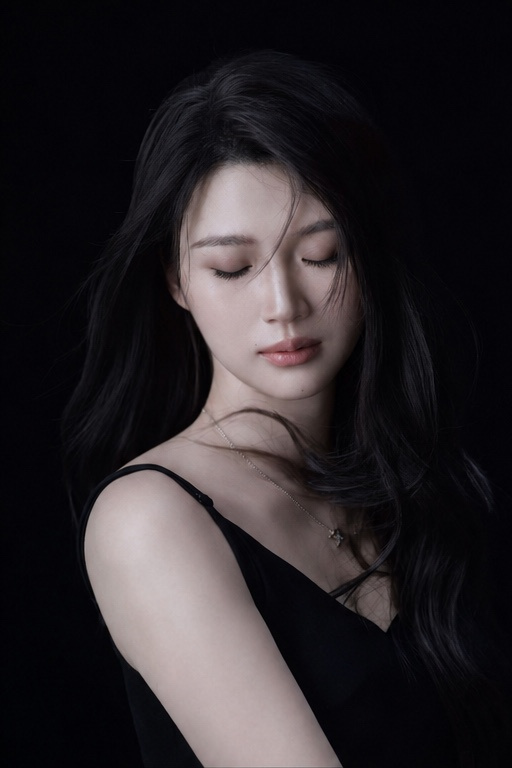 一位年轻成年东亚女性站在纯黑色摄影棚背景前，画面为近景到半身肖像，人物位于中央略偏右。身体以三分之二侧身朝向画面左侧，左肩更靠近镜头，头部轻轻回...