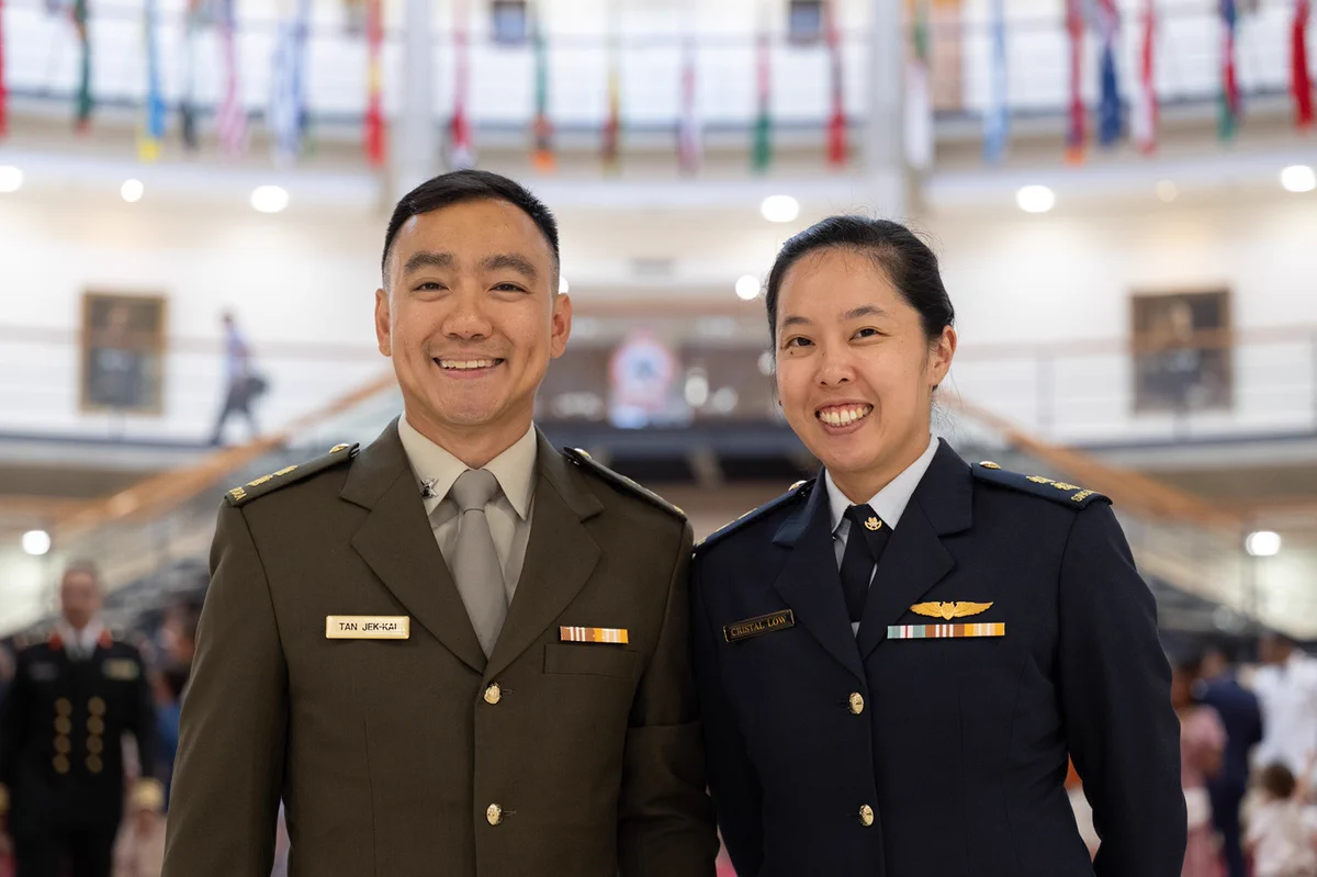 Two smiling military officers in uniform standing side by side inside a large building with flags hanging above.