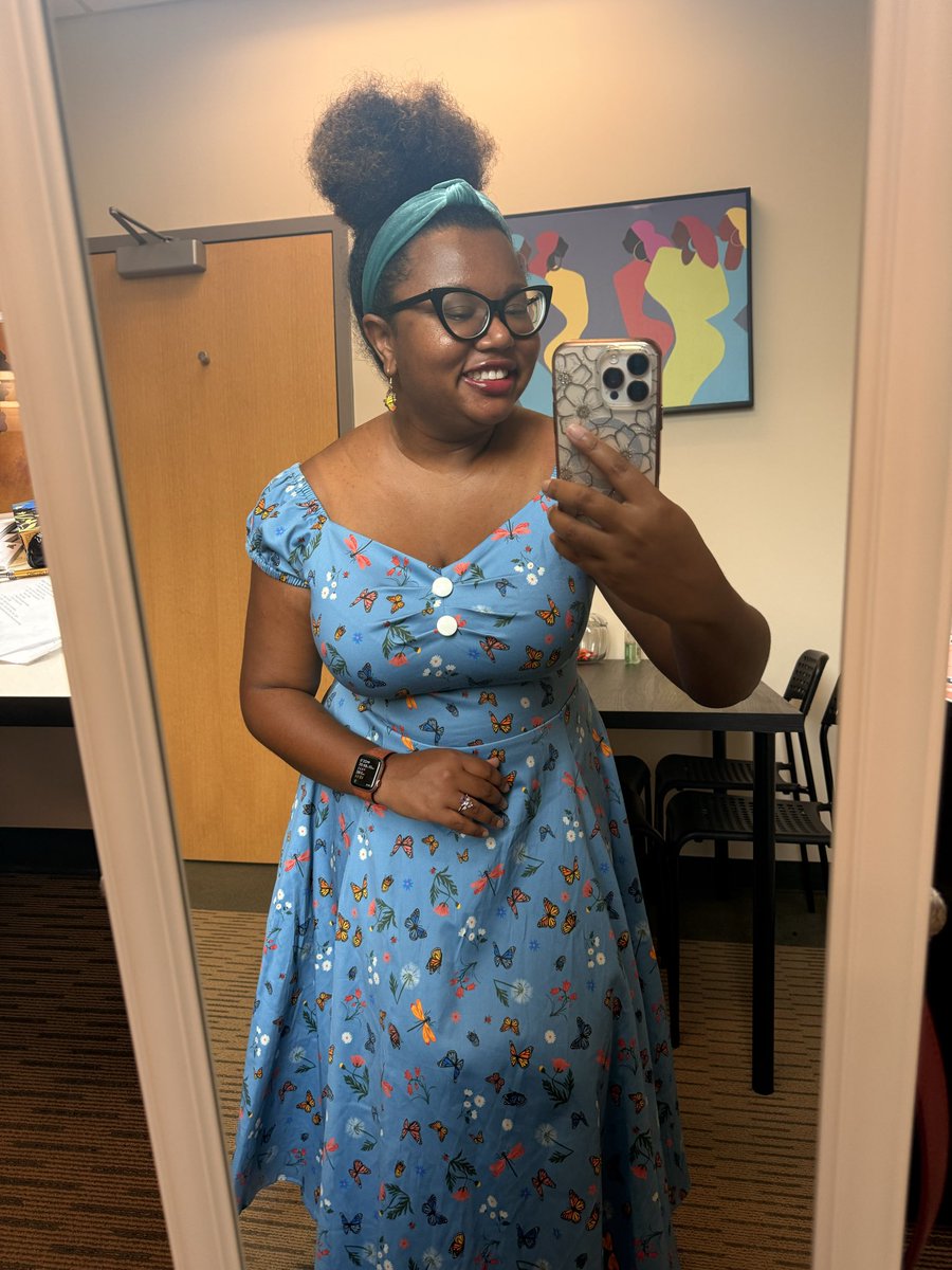 A Black woman professor taking a full-body selfie in a mirror in her office. She’s wearing a light blue dress covered in butterflies, dragonflies, and flowers with blue headband. She has black cat eye glasses and her hair is in an Afro puff.