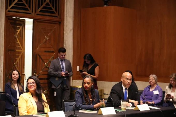 Congresswoman Summer Lee sits with other members at the Earth Justice roundtable. She is wearing a navy blue blazer and listening attentively.