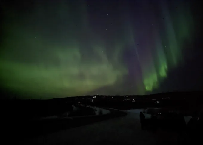 Vibrant green and purple Northern Lights paint the dark night sky with stars visible. Below, a snowy landscape stretches, featuring a winding path and distant, scattered lights from a small settlement.