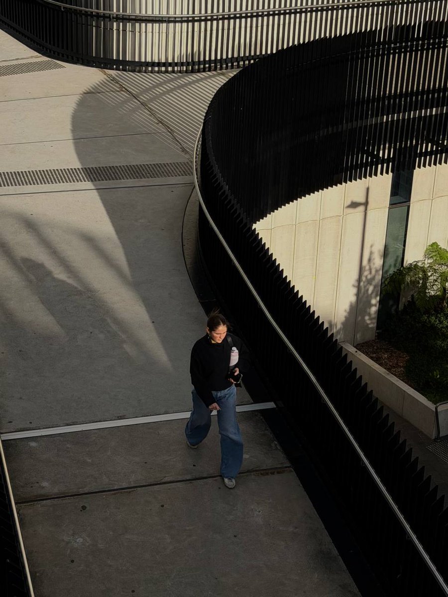 EeShayne013's tweet photo. A person walks along a concrete pathway beside a curved black railing in a modern building. Long afternoon shadows stretch across the ground, while the curved architecture guides the viewer's eye toward the subject. #ALM101DC #StreetPhotography https://t.co/c7FgVfLha3