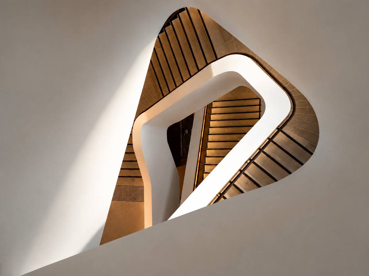 EeShayne013's tweet photo. A modern indoor staircase photographed through a curved triangular opening in a white wall. The image features repeating staircases and strong leading lines that draw the viewer's attention toward the centre of the composition.#ALM101DC #ArchitecturePhotography https://t.co/J4PV9L5DYp